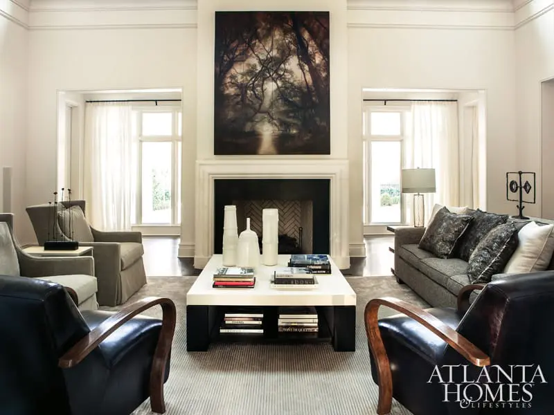 Living room with fireplace, large abstract art, neutral upholstered seating, dark leather armchairs, and a white coffee table with books and vases.