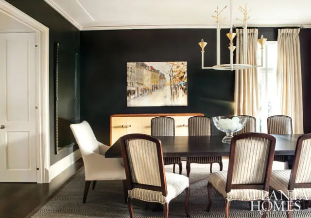 Dark wood dining table with striped upholstered chairs and a white upholstered accent chair, set against a dark wall with a street scene painting and a light wood sideboard.