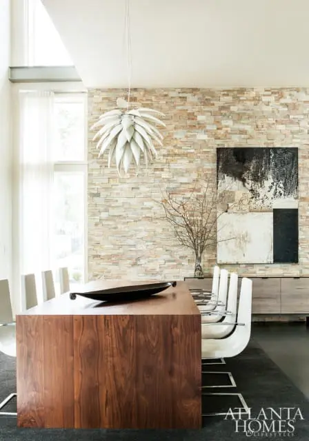 Modern dining room with a large wooden table, white chairs, a stone accent wall, and abstract art.