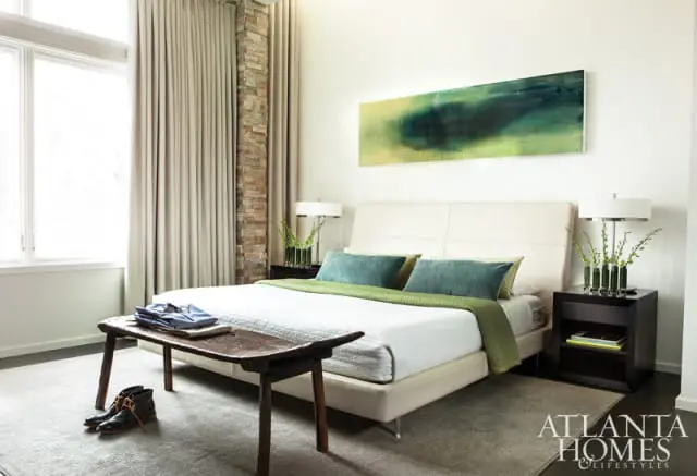 Modern bedroom with a cream upholstered bed, teal and green bedding, stone accent wall, and abstract art above the headboard.