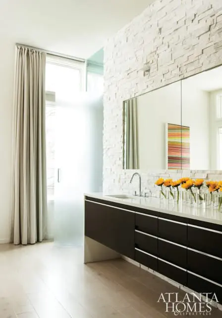 Modern bathroom with dark wood vanity, white stone accent wall, and a row of yellow flowers in glass vases.