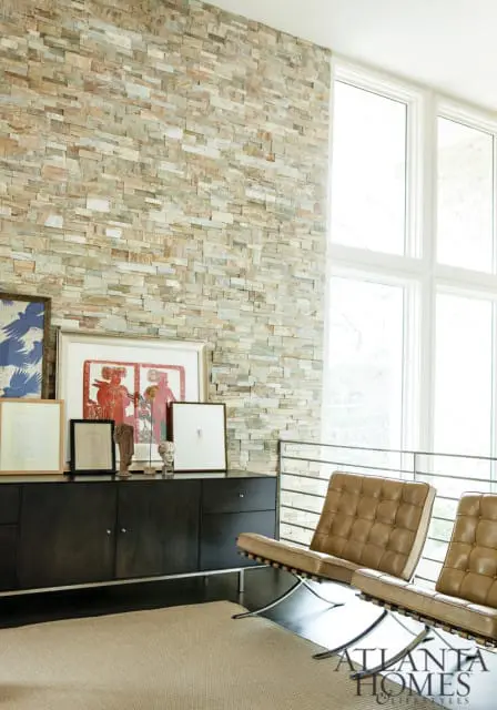 Living room with stacked stone accent wall, dark wood credenza displaying framed art and sculptures, and two tan leather Barcelona chairs.