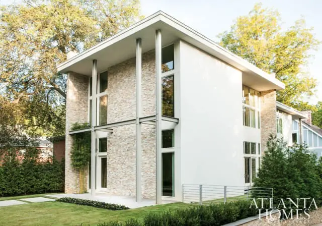 Modern two-story house with white stucco and light stone facade, tall windows, and metal columns. Lush green lawn and trees surround the property.