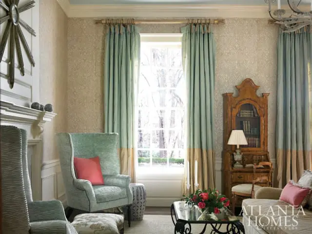 Living room with patterned wallpaper, two-tone teal and tan curtains, a fireplace, wingback chairs, and a wooden secretary desk.