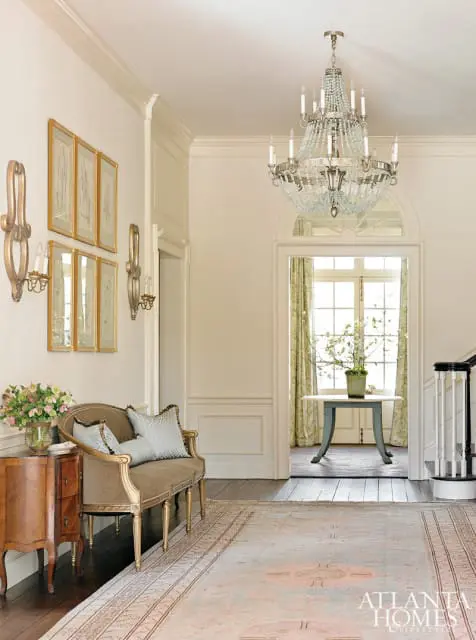 Entryway with a crystal chandelier, framed botanical prints, a settee with patterned pillows, and a Persian rug.
