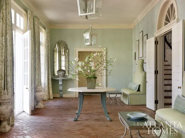 Pale green hallway with brick flooring, a hexagonal table with a floral arrangement, a tall upholstered bench, and double doors leading to a staircase.