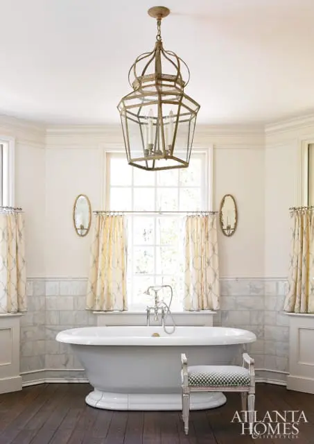 Cream bathroom with a white freestanding bathtub, marble tile wainscoting, patterned curtains, and a large ornate brass lantern chandelier.