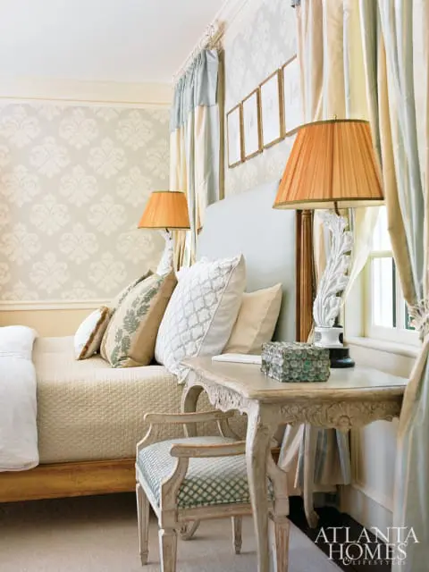 Bedroom with damask wallpaper, a light blue headboard, quilted bedspread, and ornate wooden desk with a matching chair.