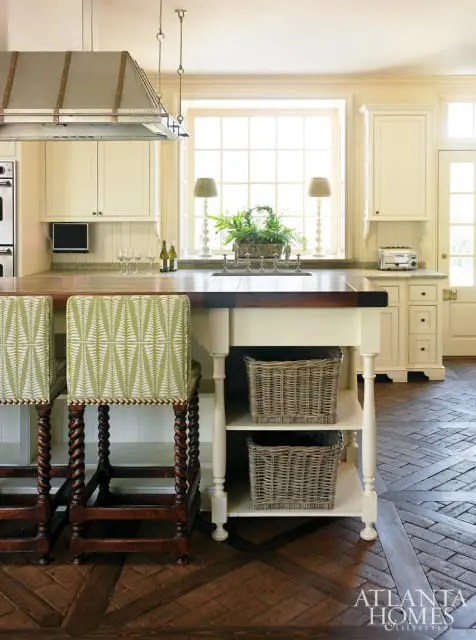 Cream kitchen with a large window, dark wood island with woven baskets, and green patterned bar stools on a brick floor.