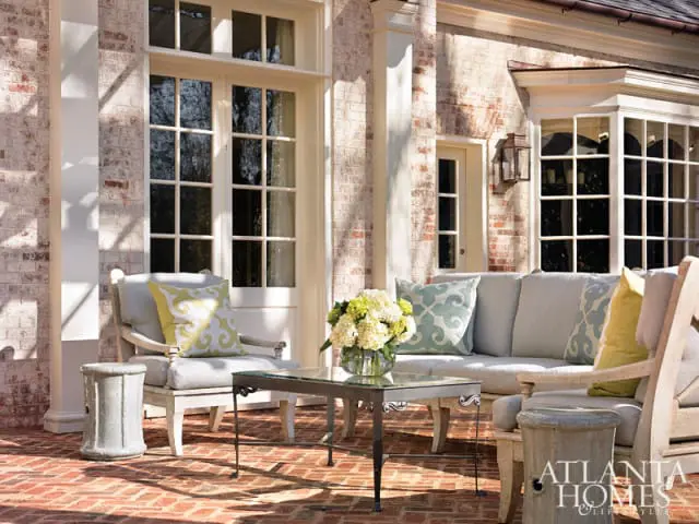 Patio with light gray upholstered seating, a glass-top coffee table, and decorative planters against a brick house with large windows.