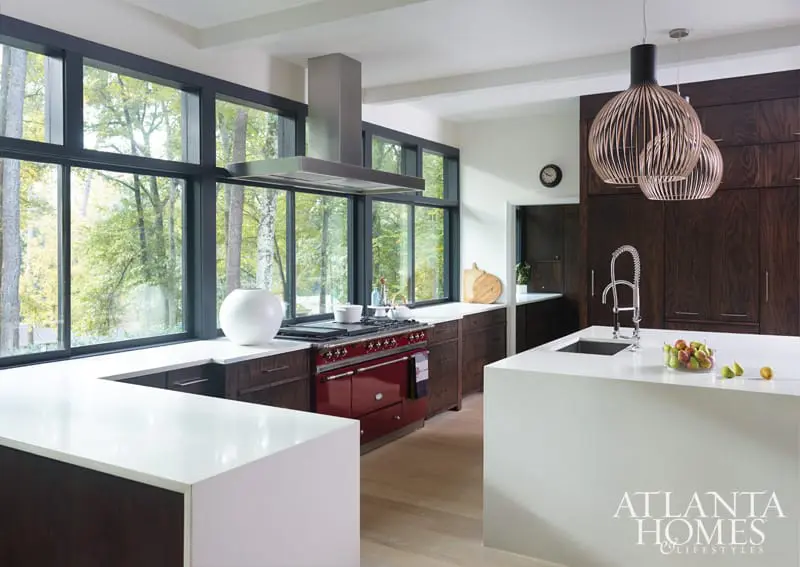Modern kitchen with dark wood cabinetry, white quartz countertops, a red range, and large windows overlooking a wooded area.