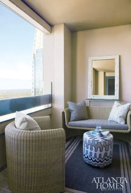 Wicker armchair and loveseat on a striped rug with a patterned ottoman and a mirror on the wall. A large window overlooks a cityscape.