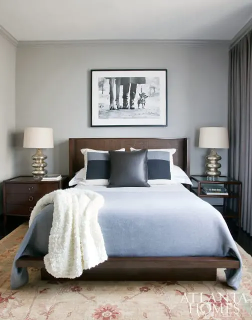 Bedroom with gray walls, a dark wood bed frame, light blue bedding, and a cream textured throw blanket. A black and white framed photograph of legs and a dog hangs above the bed.