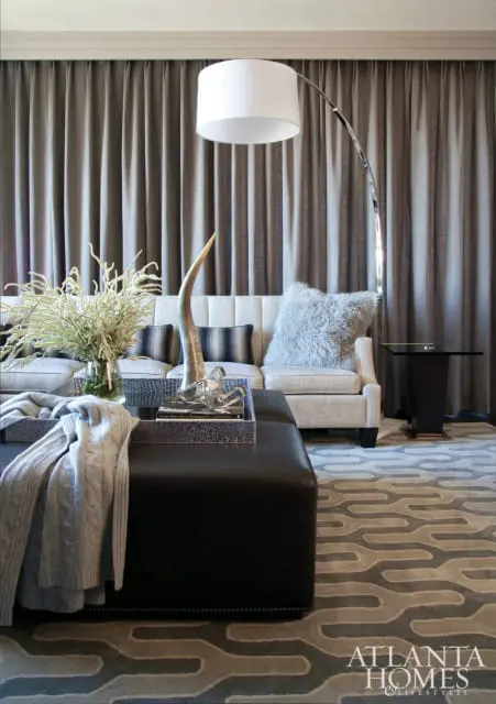 Living room with a tufted sofa, dark leather ottoman, and a modern arc floor lamp against grey floor-to-ceiling curtains.