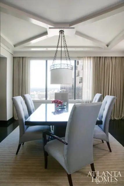 Dining room with upholstered chairs around a dark wood table with a glass top, beneath a coffered ceiling and a drum pendant light.