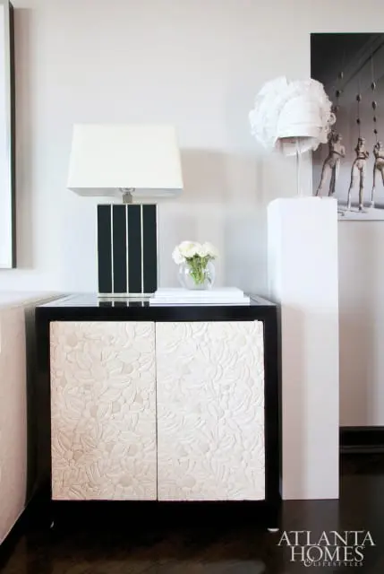 Black cabinet with carved floral doors, topped with a black and white striped lamp and a glass vase of white roses.