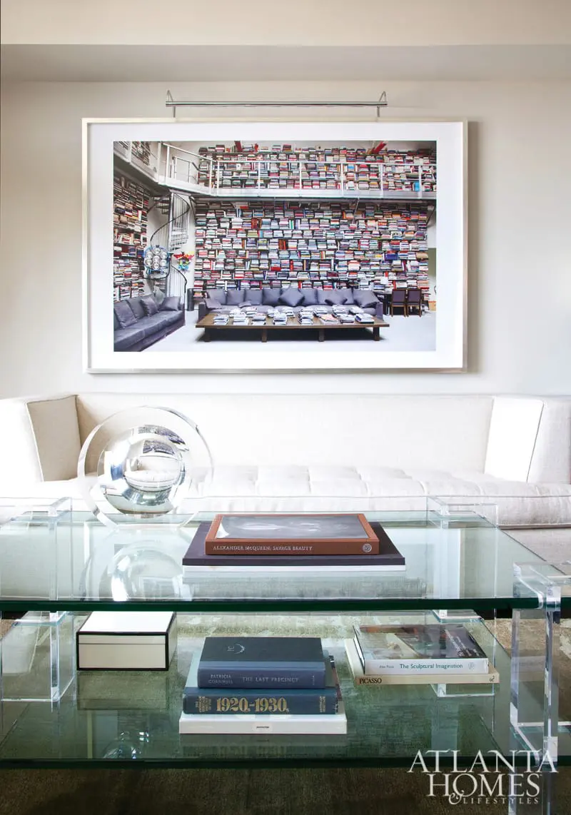 Living room with a large framed photograph of a library on the wall, a white sofa, and a glass coffee table displaying books and decorative objects.