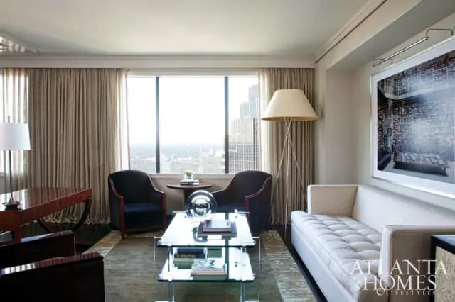 Living room with tufted cream sofa, glass coffee table, dark upholstered armchairs, and a large framed artwork above the sofa. Floor-to-ceiling curtains frame a city view.