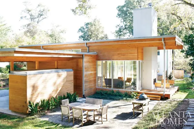 Modern home with wood siding, large windows, and a white chimney. Outdoor patio furniture and a grill are visible on the grounds.