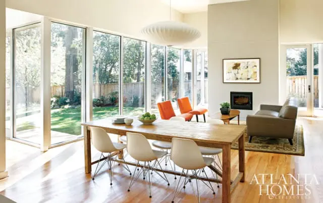 Dining area with a long wooden table and white chairs, facing large windows overlooking a backyard. A modern fireplace and leather sofa are visible in the background.