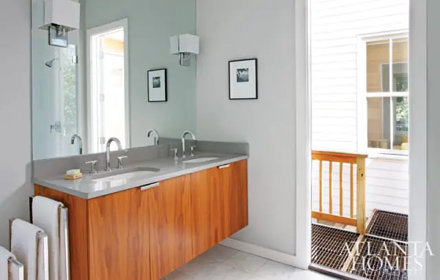 Modern bathroom with a floating walnut vanity, dual sinks, and a large mirror. Light gray walls and marble tile flooring. A doorway opens to a white-sided exterior with a wooden railing.