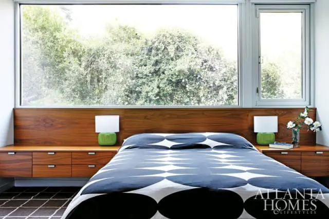 Bedroom with a large horizontal window overlooking trees, featuring a walnut headboard with built-in nightstands, a bed with a blue and white geometric pattern duvet, and green lamps.