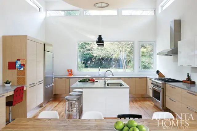 Modern white kitchen with light wood cabinetry, stainless steel appliances, and a large island with a sink. High ceilings with clerestory windows overlook a wooded backyard.