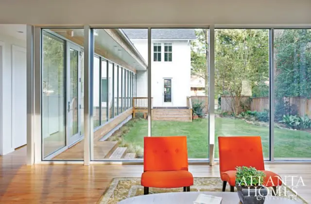 Living room with two orange accent chairs overlooking a backyard through floor-to-ceiling windows, featuring a wooden deck and a white house