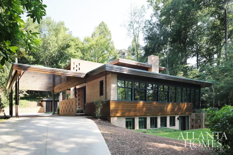Modern brick house with wood accents, large windows, and a covered carport, nestled among trees.