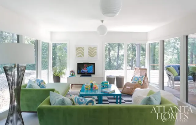 Living room with lime green sofas, a turquoise coffee table, striped rug, wicker chairs, and large windows overlooking a wooded area.