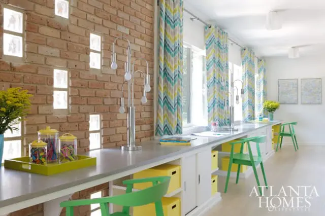 Craft room with a long grey countertop, exposed brick wall with glass block windows, and chevron curtains in yellow, blue, and teal. Green chairs and yellow storage bins add pops of color.