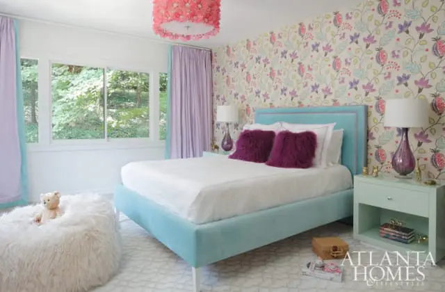 Turquoise upholstered bed with white linens and purple furry pillows, next to a mint green nightstand and a fluffy white beanbag chair. Floral wallpaper and lavender curtains frame the room.