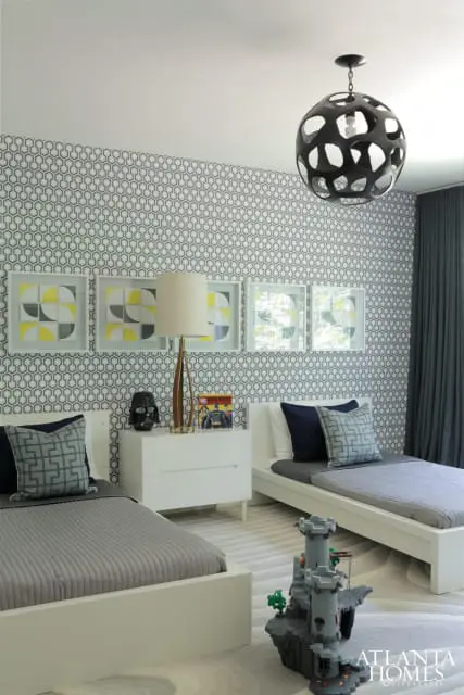 Modern bedroom with two white twin beds, gray bedding, geometric wallpaper, and a black orb pendant light. A white nightstand holds a lamp and comic books.