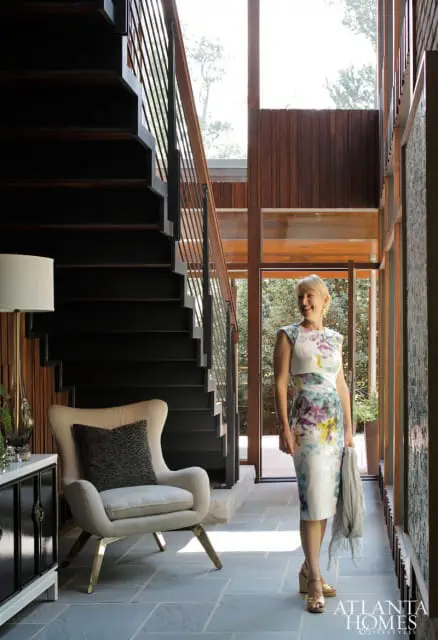 Woman in floral dress standing by a modern staircase with dark wood treads and metal railings, next to a cream armchair and a black cabinet.