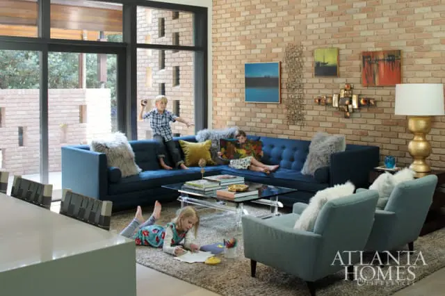 Living room with exposed brick walls, featuring a deep blue sectional sofa, light blue armchairs, a glass and acrylic coffee table, and children playing.