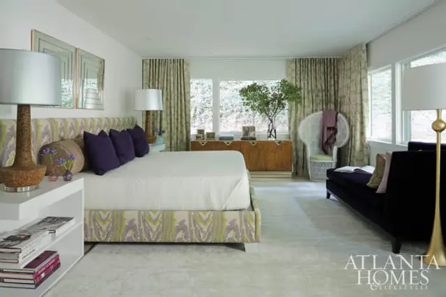 Bedroom with a patterned headboard and bed skirt in green and purple, white bedding, purple accent pillows, and a white nightstand with books.