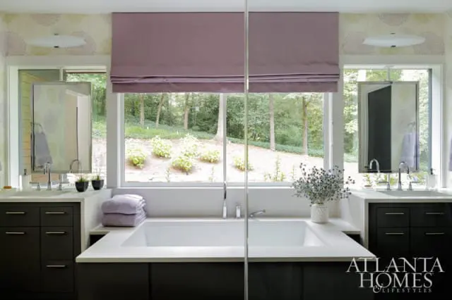 Modern bathroom with a freestanding bathtub, dual vanities with dark cabinetry, and a large window overlooking a wooded landscape.