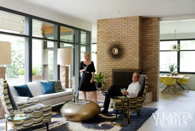 Living room with a white sofa, patterned armchairs, a large brass coffee table, and a brick fireplace. A man and woman stand and sit, holding cocktails.