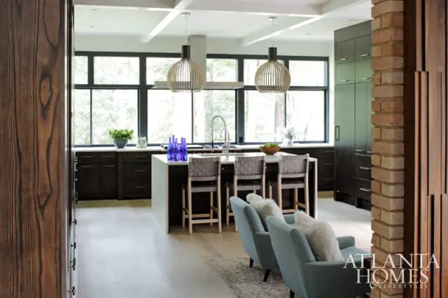 Modern kitchen with dark wood cabinetry, white island with bar stools, and large windows overlooking trees. Two pendant lights hang above the island.