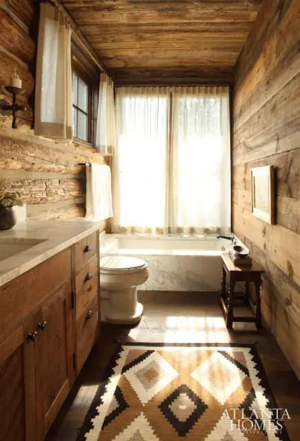 Rustic bathroom with log walls, wooden vanity with marble countertop, white toilet, marble-clad bathtub, and a geometric patterned rug.