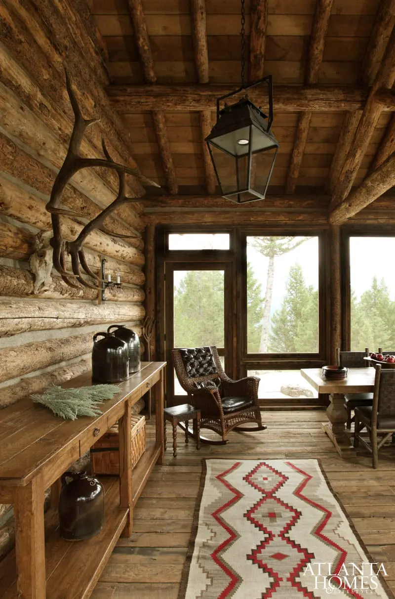 Log cabin interior with wood-paneled walls and ceiling, a large antler display, a wicker rocking chair, and a dining table by a window overlooking a forest.