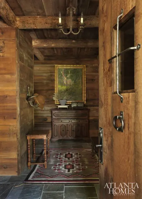 Log cabin entryway with wood-paneled walls, a painting of a deer, antique wooden furniture, a patterned rug, and a wrought iron chandelier.