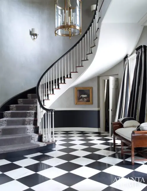 Grand foyer with a curved staircase, black and white checkered marble floor, gray textured walls, and black and white striped curtains.