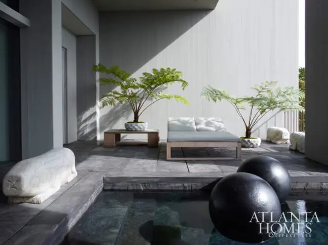 Modern patio with gray stone flooring, a light wood chaise lounge, a low wood table with a potted fern, and abstract sculptures.