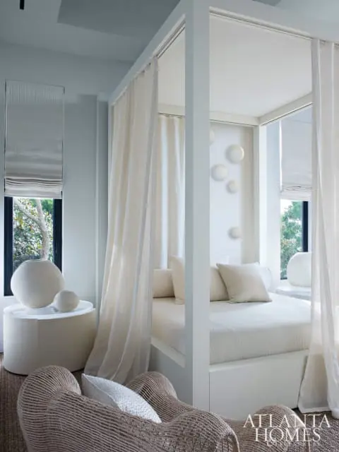 Bedroom with a white four-poster bed draped in sheer curtains, a woven armchair, and decorative round wall art.