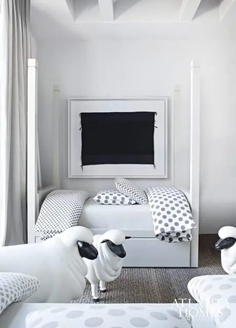 White bedroom with a four-poster bed, grey and white polka dot bedding, sheep-shaped stools, and framed abstract art on the wall.