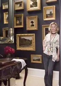 Woman in floral blouse and jeans leans against a dark wall adorned with framed landscape paintings. A vanity with a sink and red flowers is in the foreground.