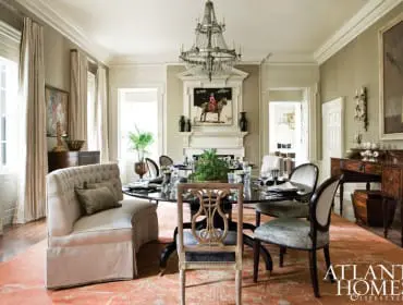 Formal dining room with a large oval table set for dinner, tufted banquette seating, ornate chairs, a crystal chandelier, and a fireplace with artwork above. A large coral rug covers the hardwood floor.