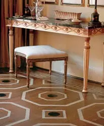 Ornate wooden console table with marble top and upholstered stool in front of a wall with draped curtains. The floor features a geometric inlay pattern of wood and stone.