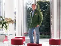 Man in green blazer and jeans standing on red ottomans in a bright, modern interior with large windows overlooking greenery.